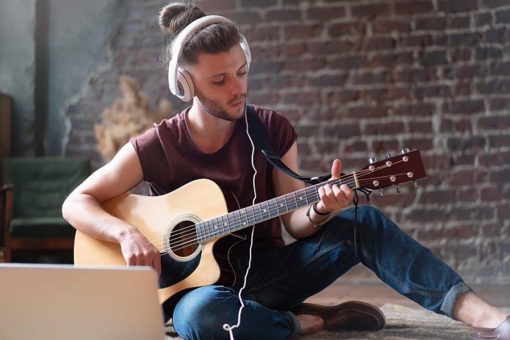 בחור עם אוזניות מחזיק גיטרה יושב על הרצפה מול מחשב נייד ומנגן