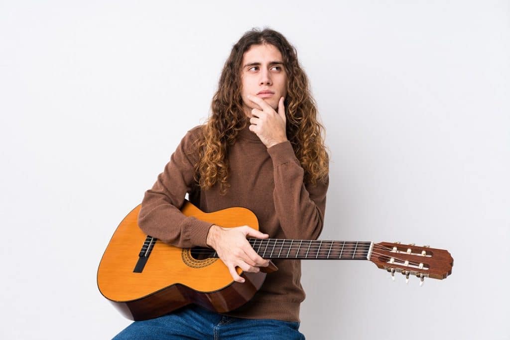 בחור עם שיער ארוך יושב וחושב עם יד על הסנטר מחזיק גיטרה רקע לבן