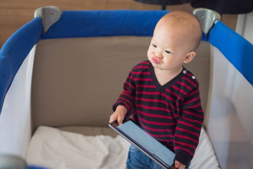 תינוק אסיאתי מחזיק טאבלט מחייך ועומד בתוך לול קמפינג כחול