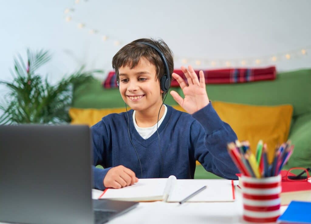ילד עם אוזניות יושב מול מחשב נייד מחייך ומנוף לומד עם מחברת