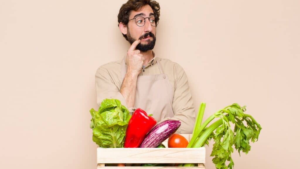 בחור עם משקפיים חושב ומסתכל לצד ולפניו ארגז עם ירקות רקע בז