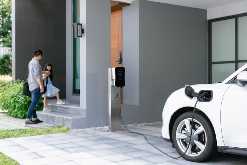 רכב חשמלי מחובר לעמדת טעינה ליד בית עם אבא וילדה