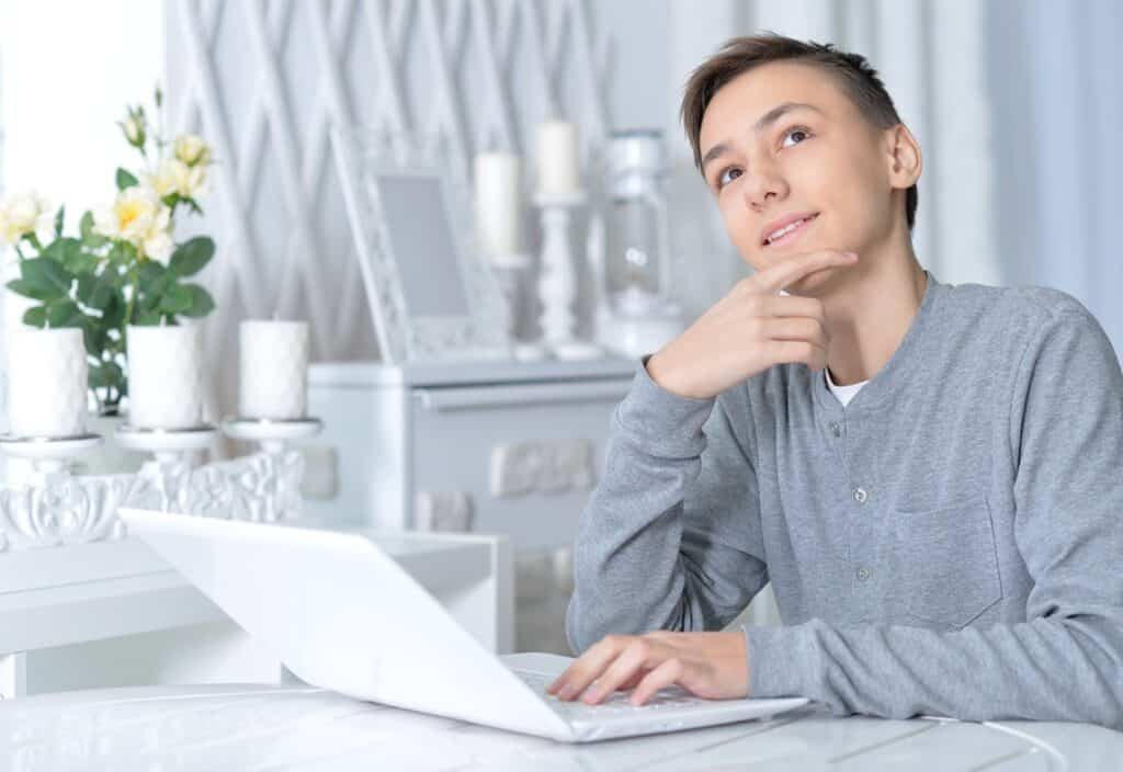 בחור בסוודר אפור בהיר יושבת וחושב עם יד על הסנטר מול מחשב נייד רקע לבן