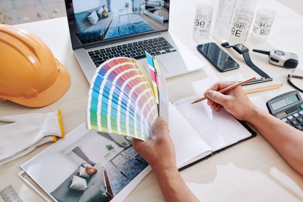 ידיים מחזיקות מניפת צבעים ועפרון מעל שולחן עבודה עם מחברת מחשב נייד ותמונות
