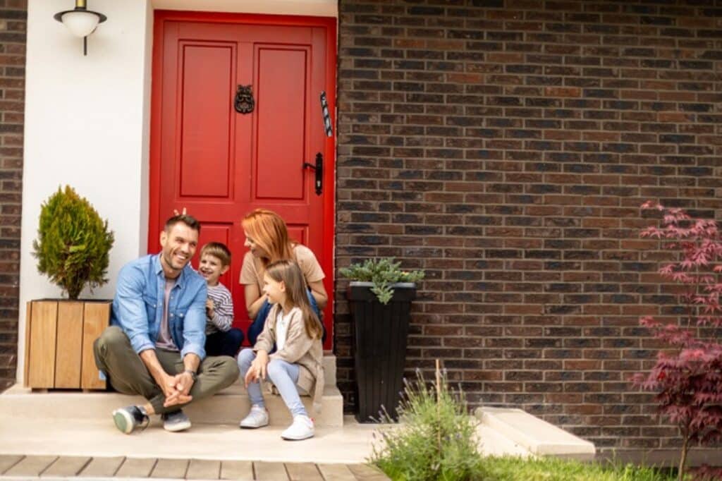 משפחה שמחה יושבת מחוץ לדלת בית הורים עם שני ילדים ליד דלת אדומה עם מזוזה