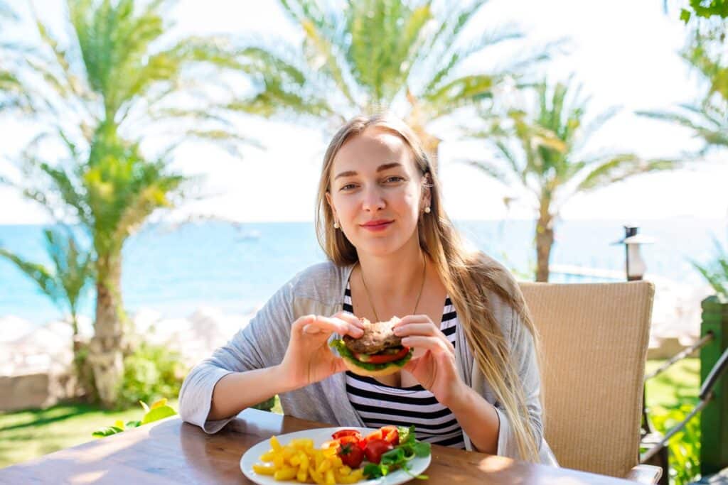 בחורה בלונדינית בחופשה מחייכת ומחזיקה המבורגר ללא גלוטן ברקע ים ועצים