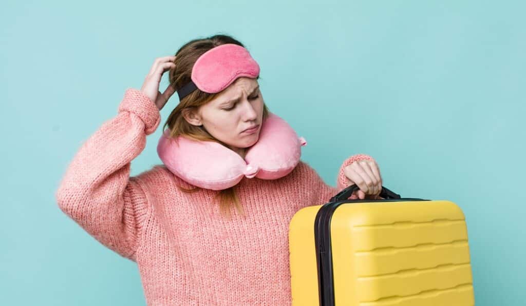 בחורה עם כרית לצוואר ומסכת שינה מחזיקה מזוודה צהובה וחושבת רקע תכלת