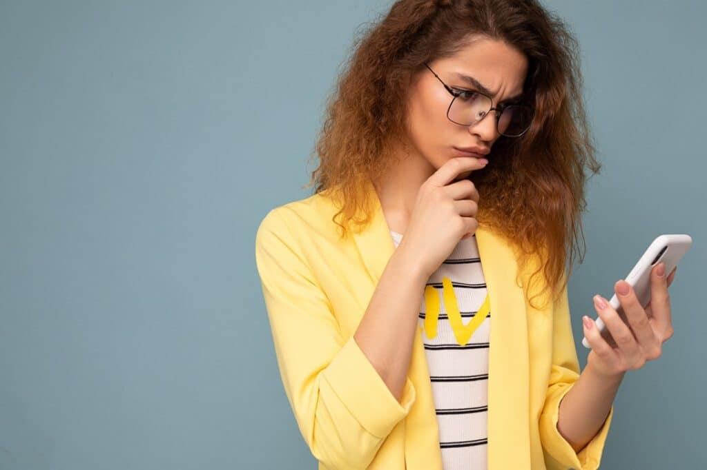 בחורה מתולתלת עם גקט צהוב ומשקפיים מחזיקה טלפון נייד וחושבת