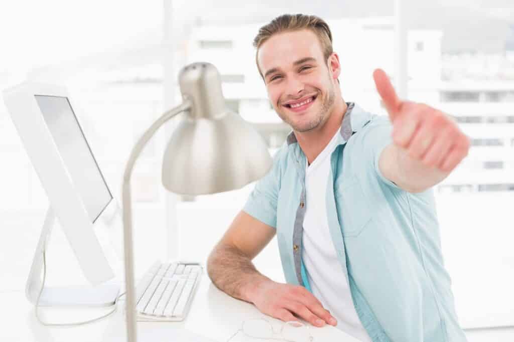בחור בחולצה מכופתרת יושב ליד מחשב במשרד ומחייך מרים אגודל