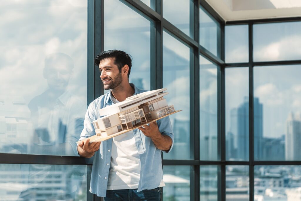 אדריכל מחזיק דגם של בית ומסתכל מהחלון ומחייך