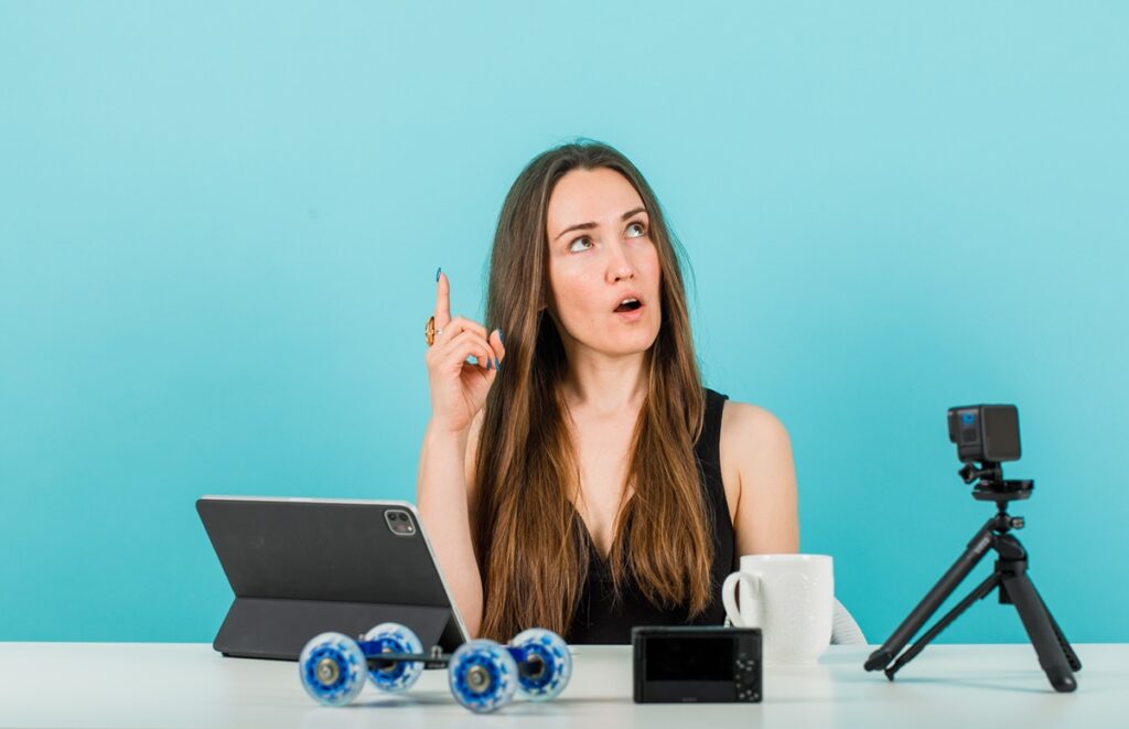 בחורה יושבת ליד שולחן עם מחשב נייד חצובה למצלמה וכוס קפה וחושבת רקע תכלת