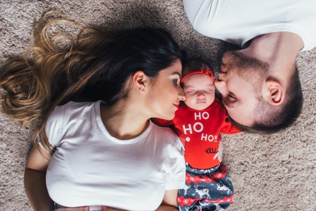 אמא ואבא שוכבים על הרצפה וביניהם תינוקת קטנה ומנשקים אותה משני הצדדים