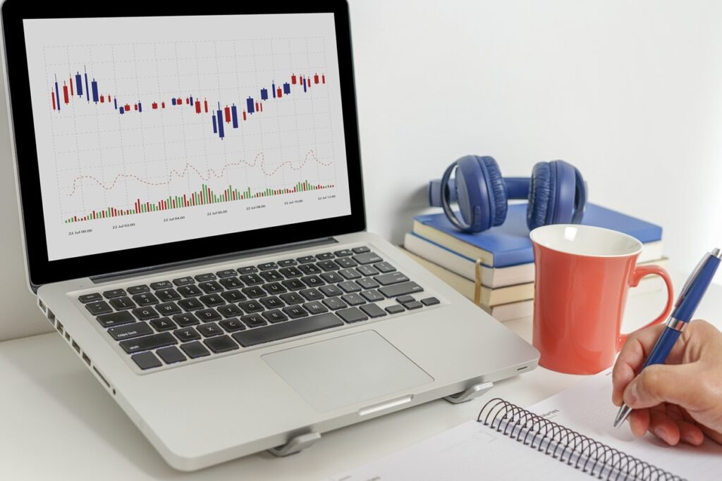 מחשב עם גרף של שוק ההון ולידו יד כותבת במחברת